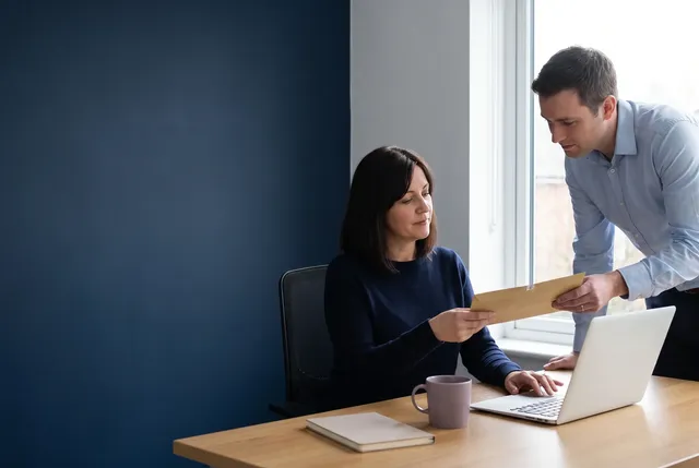 Office manager at a tidy desk receives a sealed document envelope from a colleague while sending an email on her laptop